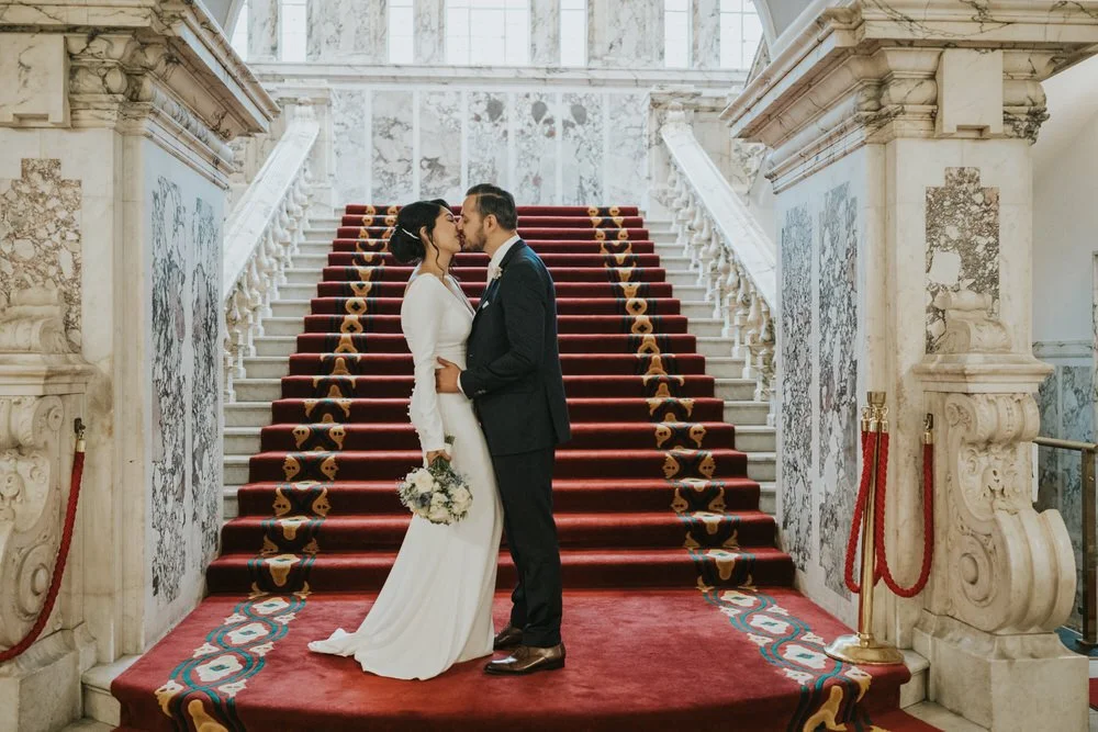 Bride and groom kissing on the red carpeted marble staircase at Belfast City Hall with red rope barriers on either side