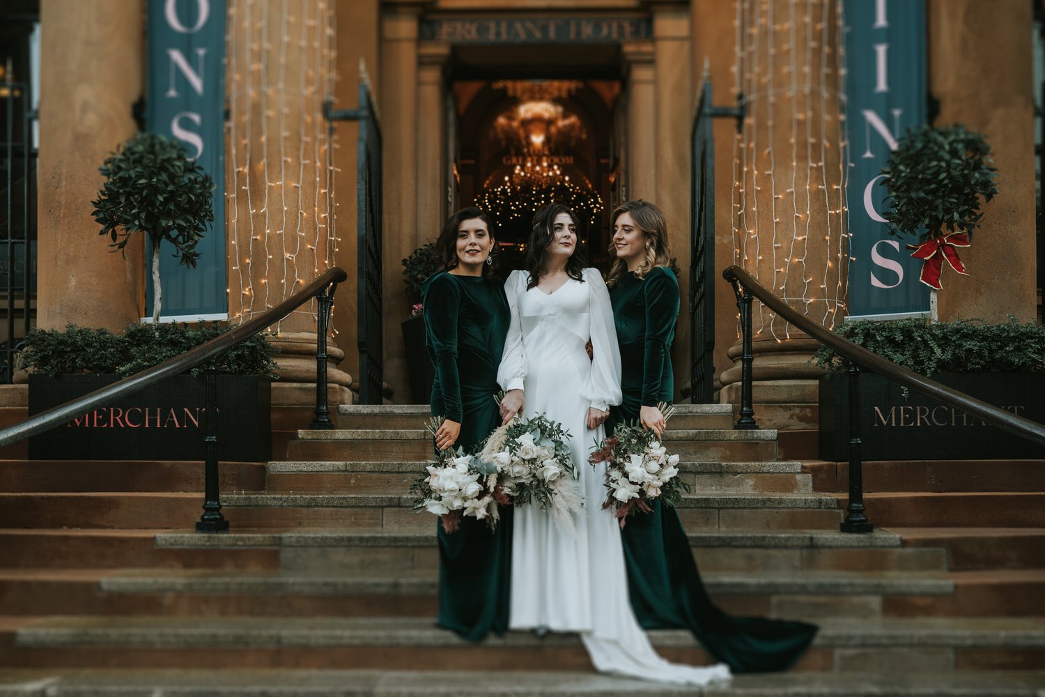 Bride standing on the front steps of The Merchant Hotel in Belfast with two bridesmaids in green velvet dresses