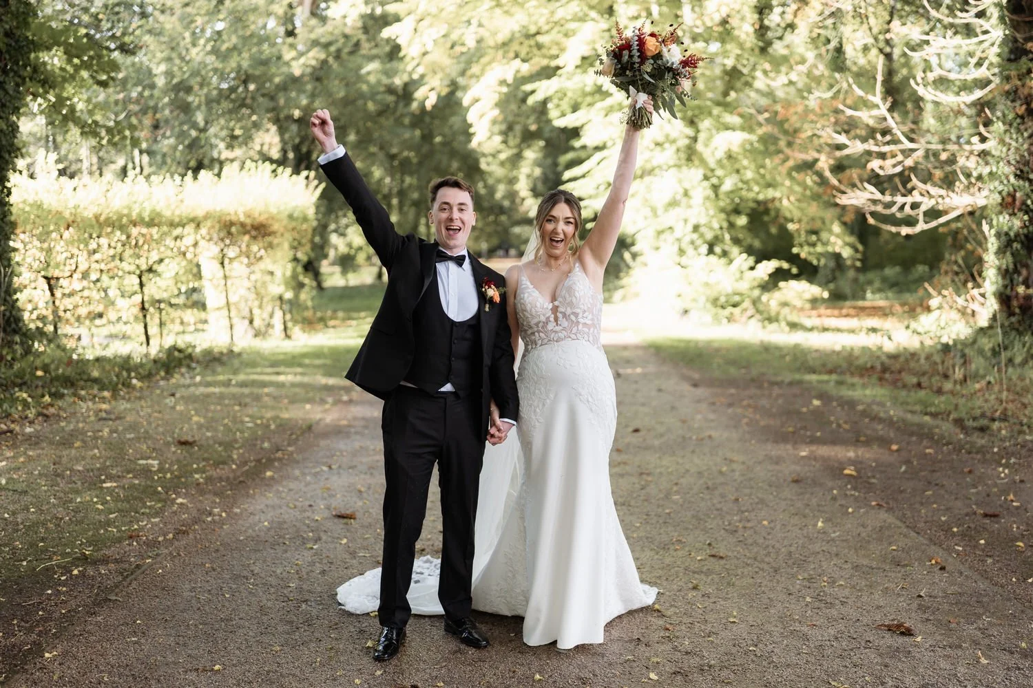 Bride and groom cheering with their arms raised on a tree lined path in Antrim Castle Gardens