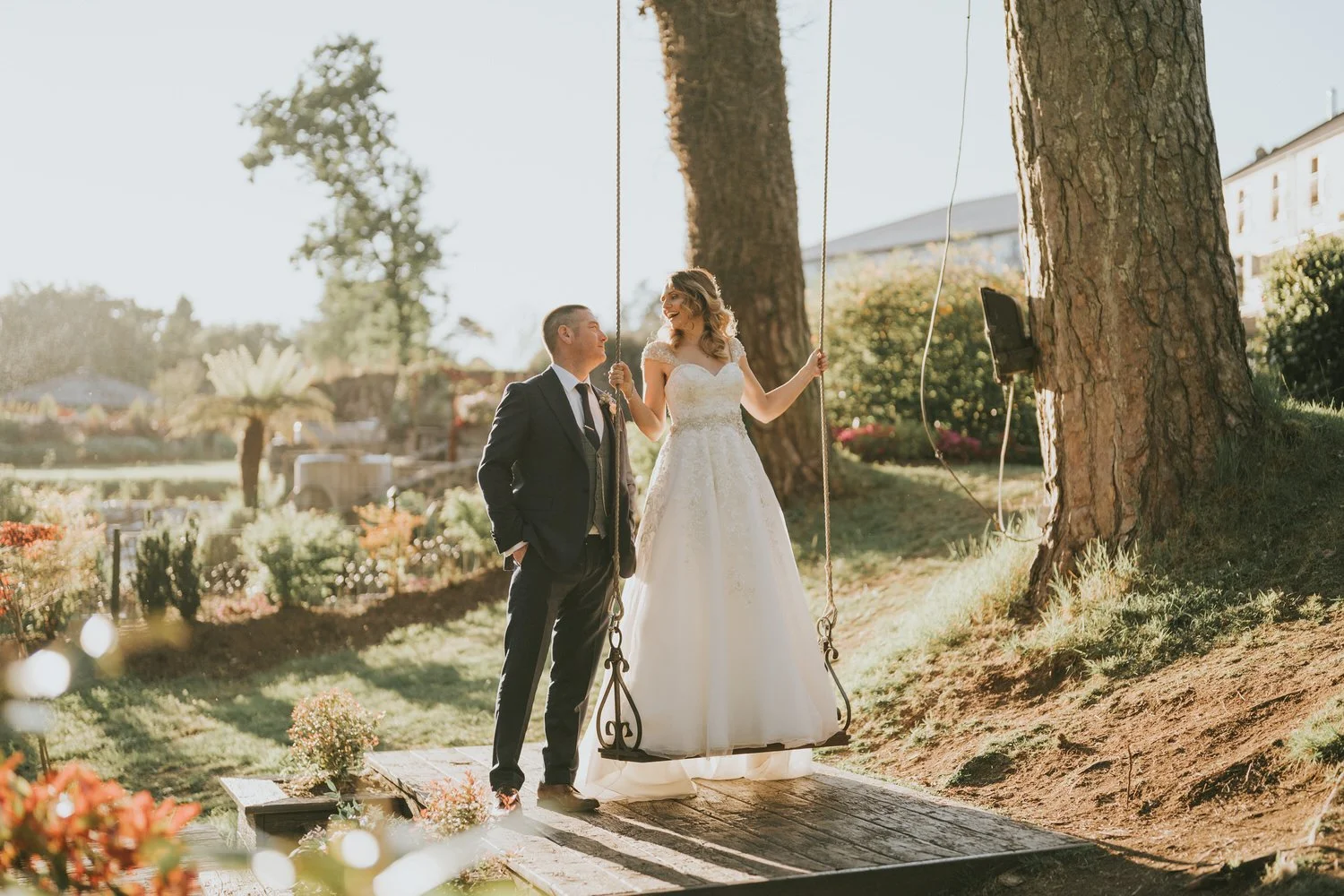 Bride and groom standing beside a wooden garden swing in the sunlit grounds at Galgorm Resort, Ballymena