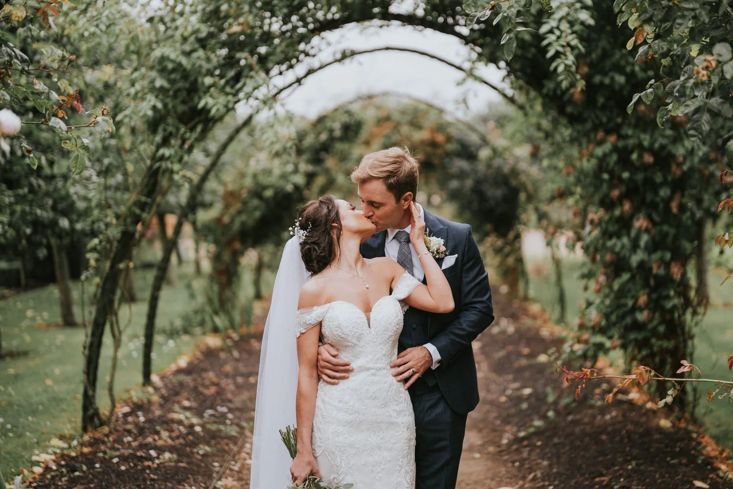 Bride and groom kissing beneath a leafy rose arch in a garden walkway