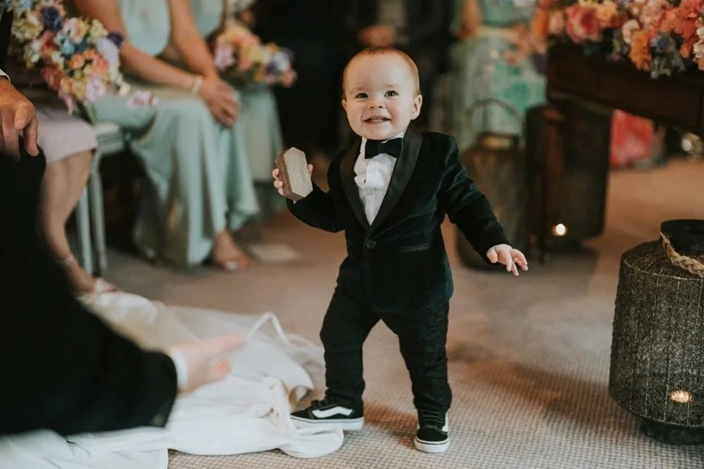 Toddler in a tuxedo smiling genuinely whilst walking at a wedding reception, a perfectly imperfect moment captured