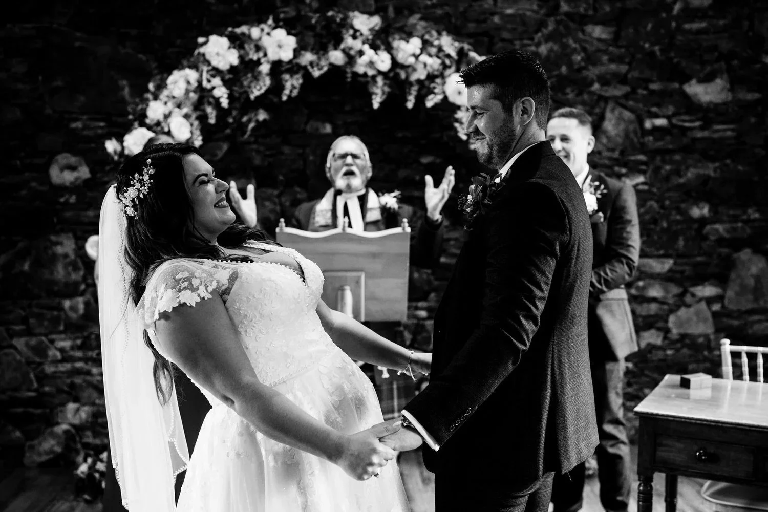 Bride and groom laughing and holding hands during their wedding ceremony in a stone chapel in Northern Ireland, captured in black and white by Pure Photo N.I