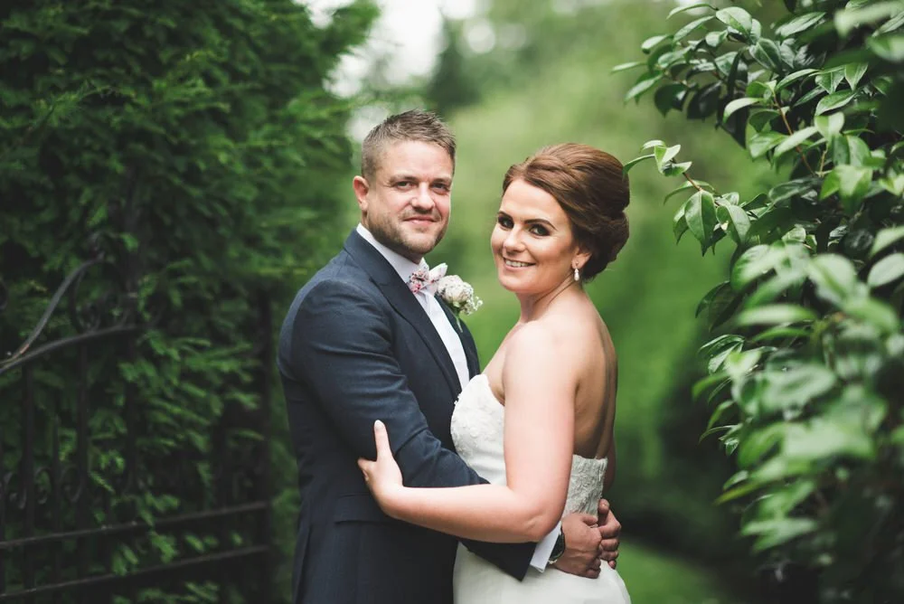 Bride and groom embracing between tall green hedges beside an old iron gate on their wedding day