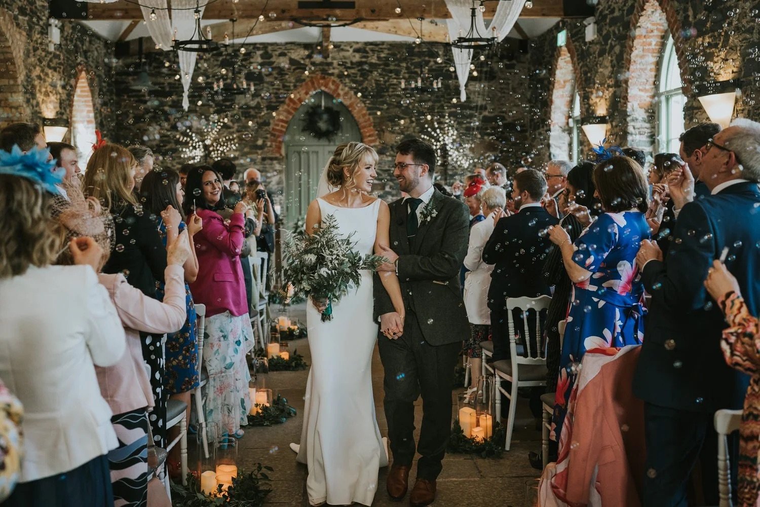 Bride and groom walking back down the aisle together surrounded by guests blowing bubbles in the stone walled main function room at Orange Tree House