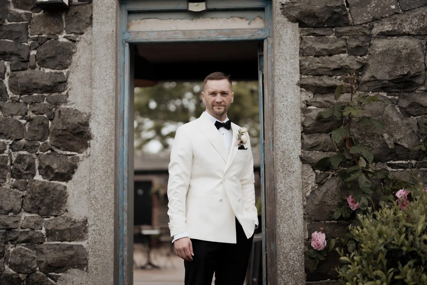Groom in a white tuxedo jacket and bow tie standing in a stone doorway on his wedding day
