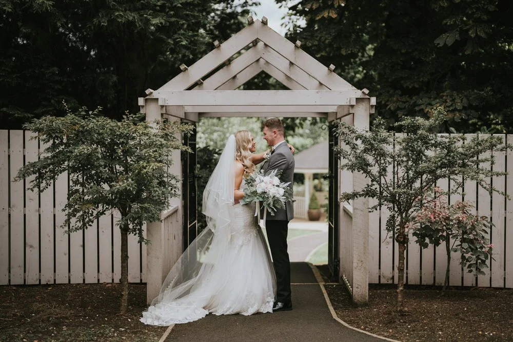 Bride and groom embracing beneath a white timber archway in the garden at Leighinmohr House Hotel, Ballymena