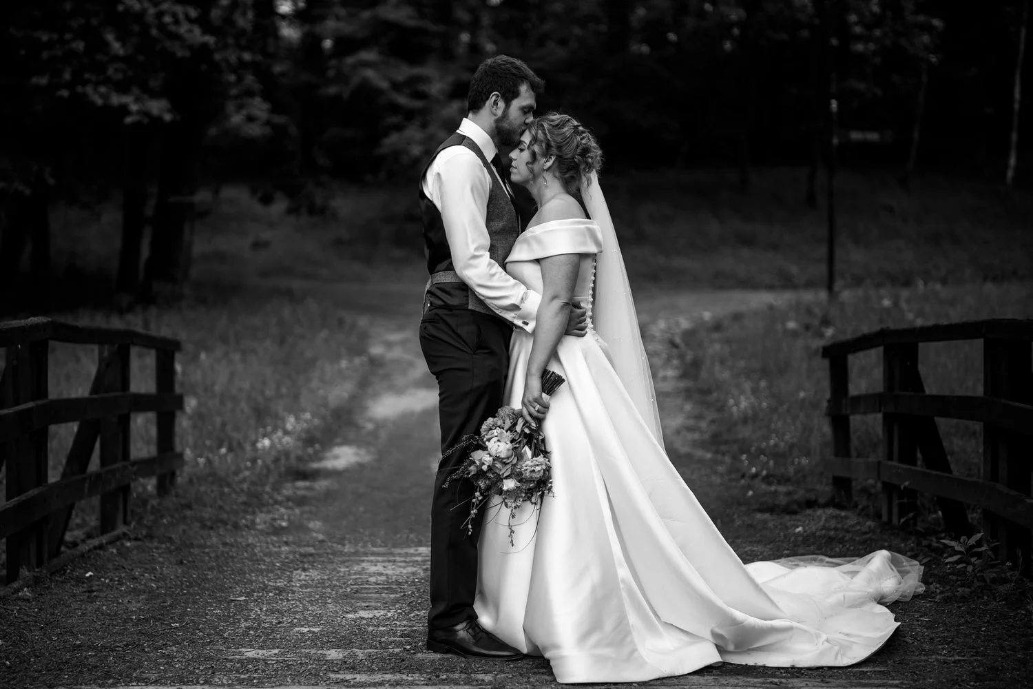 Bride and groom embracing on a woodland path beside a wooden bridge at Glenpark Estate, in black and white