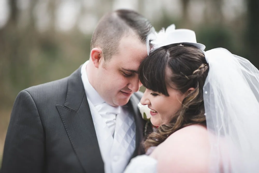 Bride in a veil and small hat resting her forehead against the groom's in the grounds of the Crowne Plaza Belfast