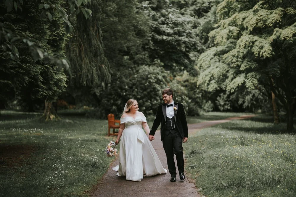 Bride and groom walking hand in hand along a tree lined garden path
