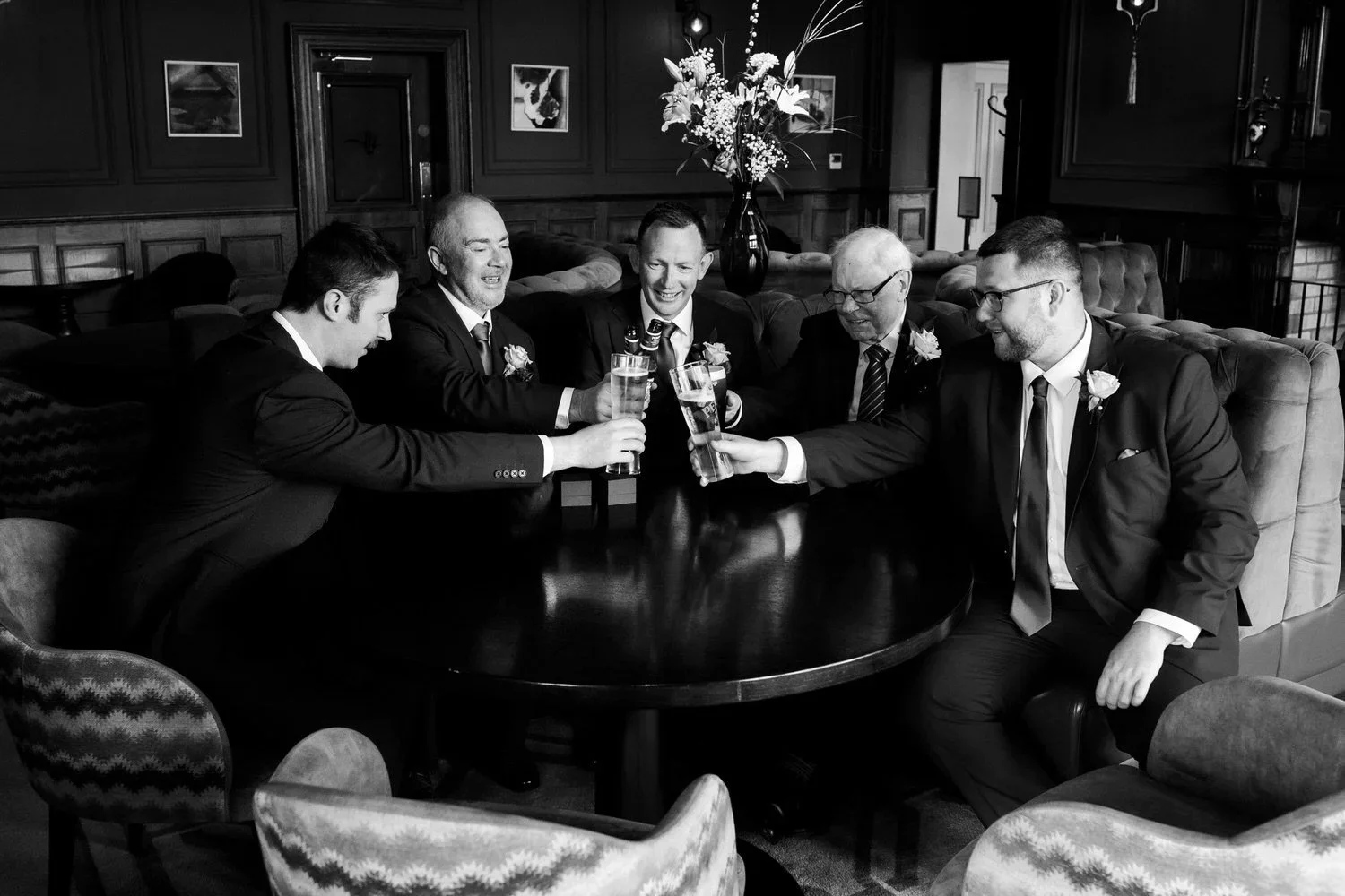 Black and white photo of a groom and his groomsmen raising a toast in a hotel lounge on the wedding morning