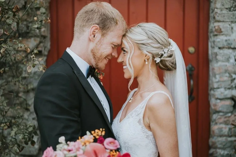A bride and groom smiling and touching foreheads in front of a red door in the grounds of Tullyglass Hotel Ballymena photographed by Pure Photo NI