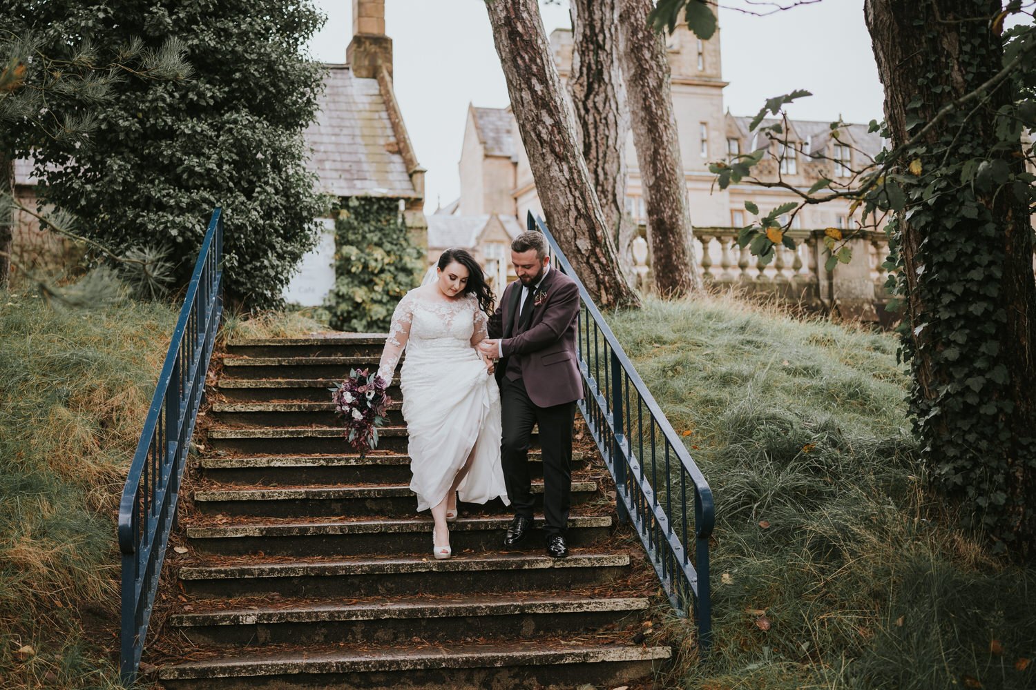 Bride and groom walking down stone steps beside a blue railing with the castle behind them at Bangor Castle