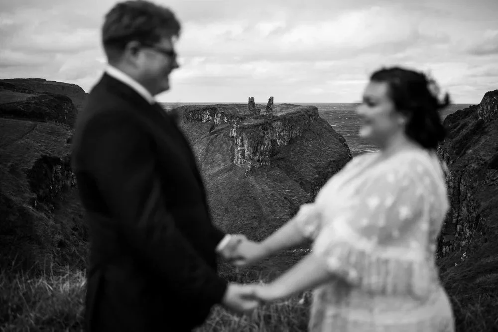 Bride and groom holding hands during a coastal elopement in Northern Ireland with Dunseverick Castle ruins visible on the clifftop behind them, photographed by Pure Photo NI