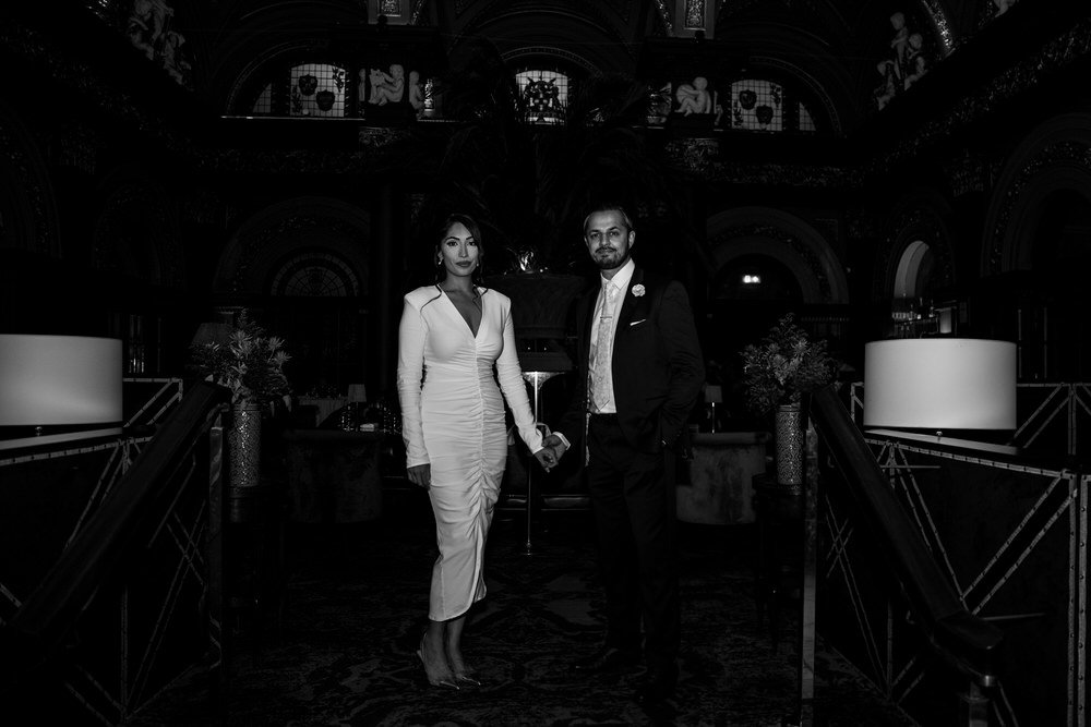 Bride and groom standing hand in hand in the grand, dimly lit interior of The Merchant Hotel in Belfast, in black and white