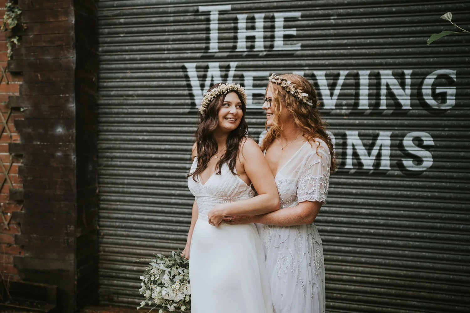Two brides in flower crowns embracing in front of a grey shutter with painted lettering at The Wool Tower