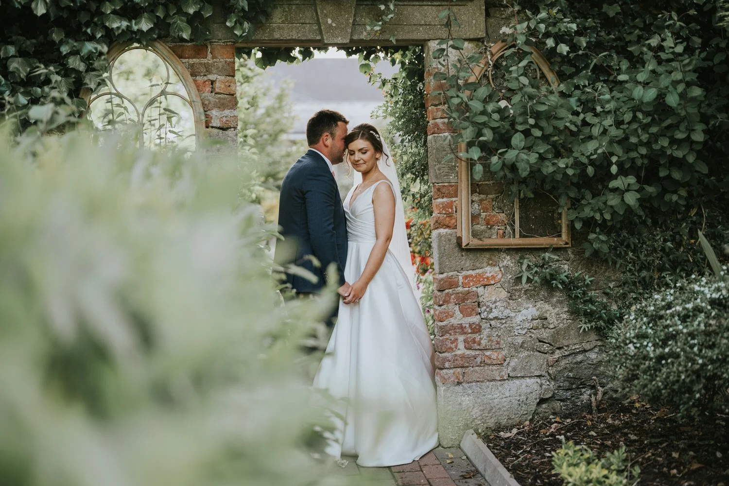 Bride and groom standing close together beside an old brick garden wall with ivy and a mirror at Corick House