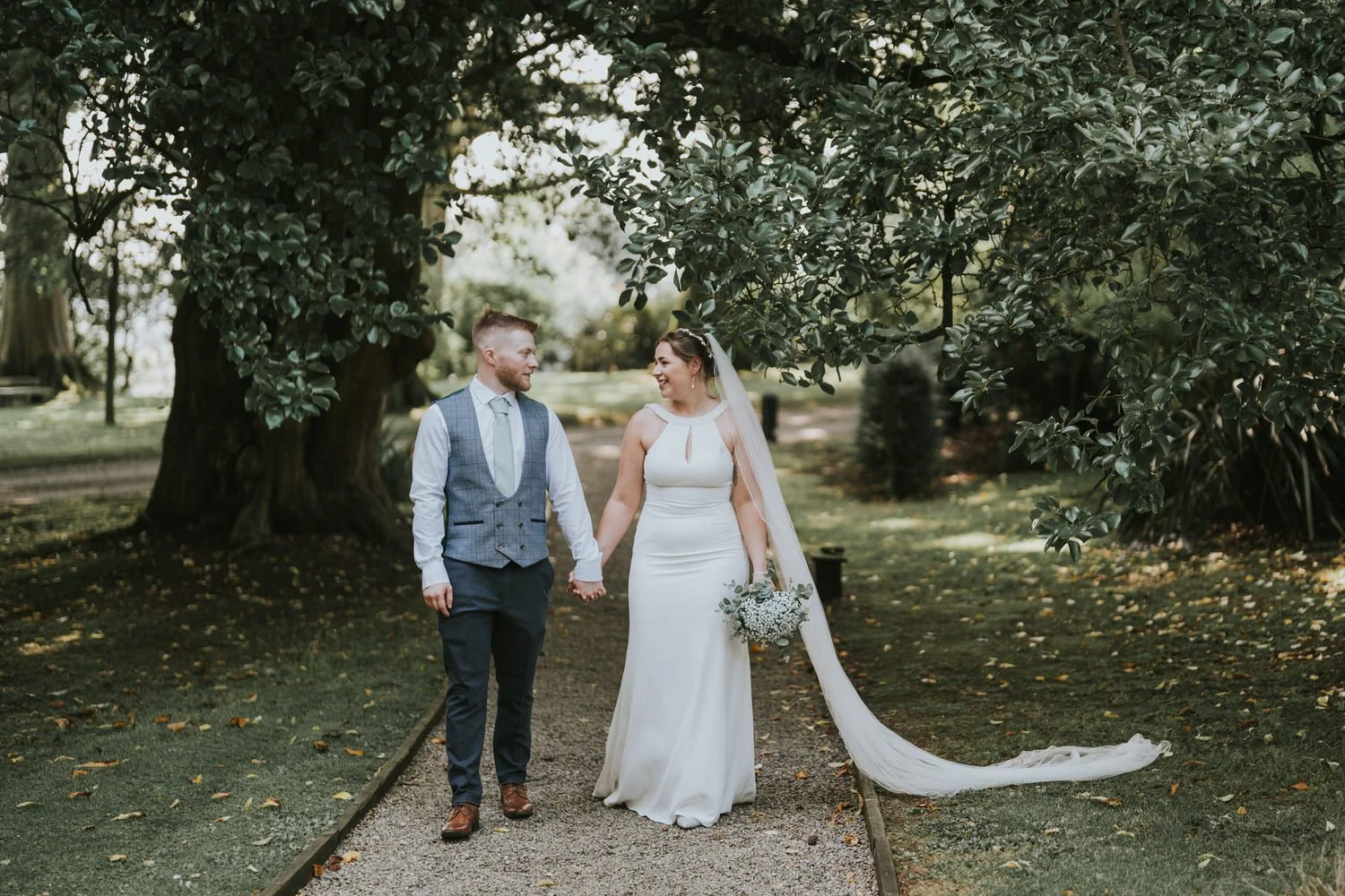 Bride and groom holding hands on a gravel path under mature trees at Tullyveery House, the bride's long veil trailing behind her