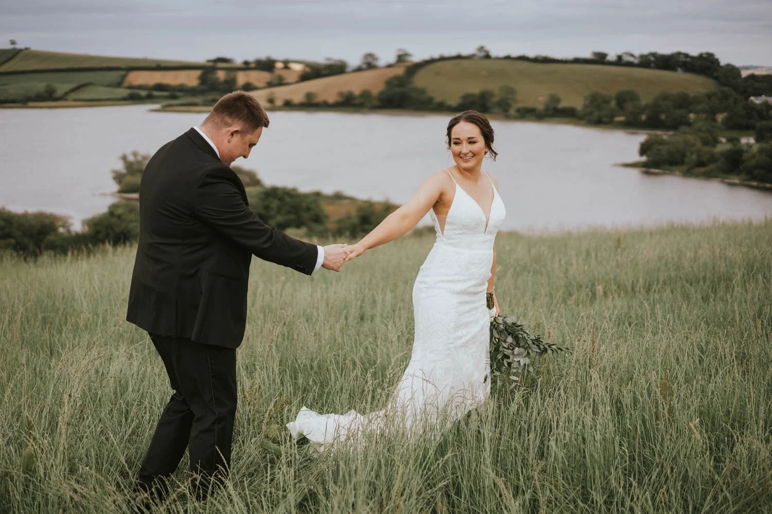 Groom leading the bride by the hand through long grass beside the water at Tullyveery House