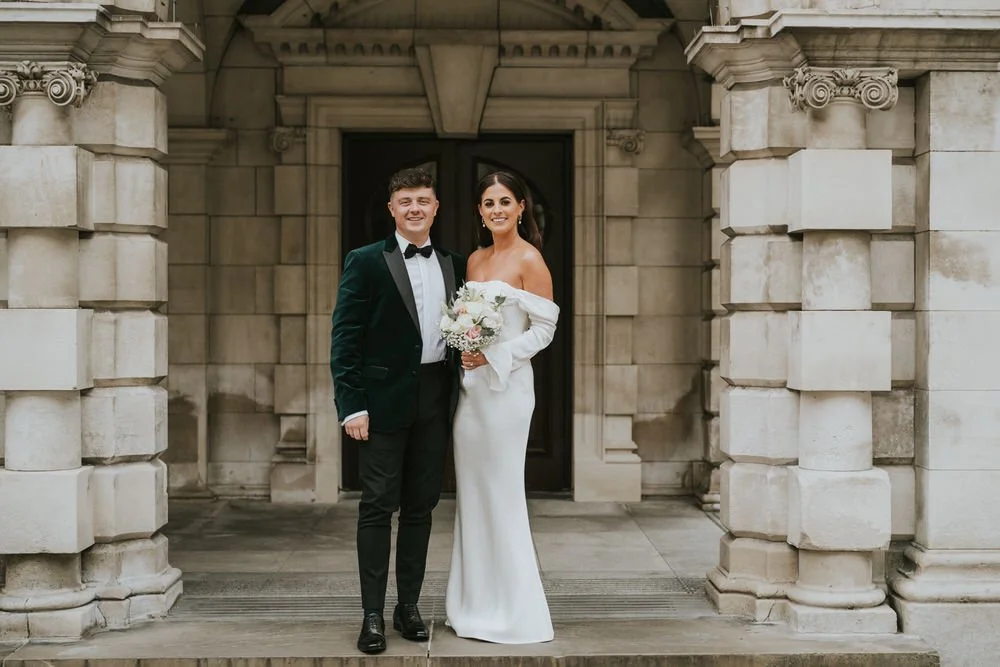 Bride in a white gown and groom in a dark velvet jacket standing in the stone doorway at Belfast City Hall