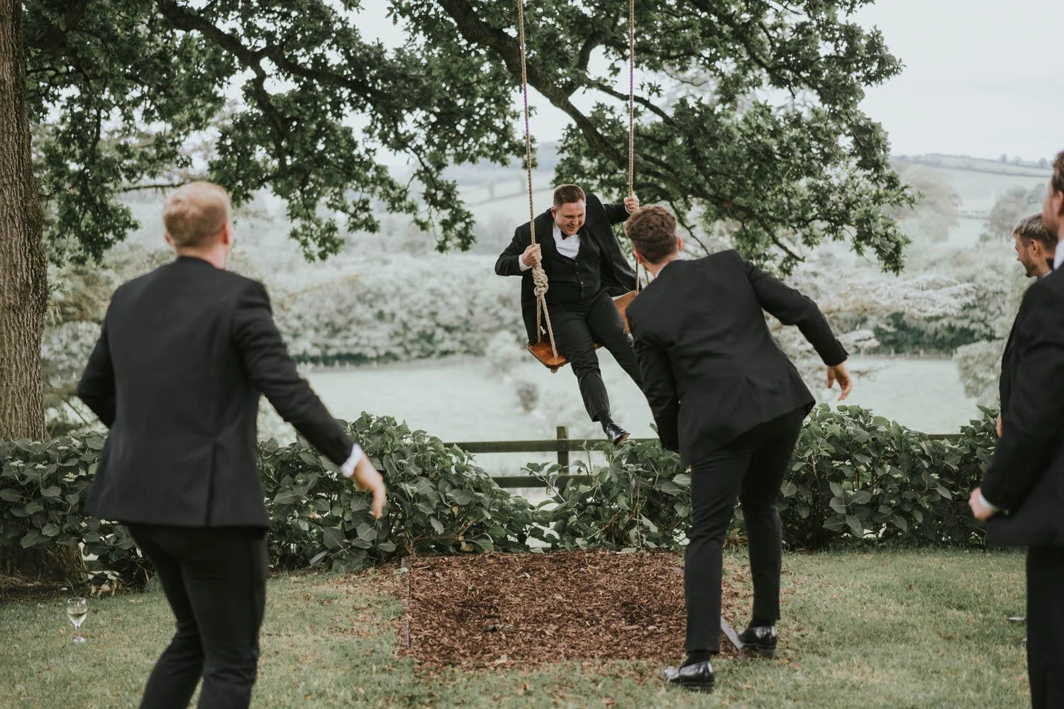 Groomsman laughing on a tree swing as friends gather round, with a view over the countryside at Tullyveery House