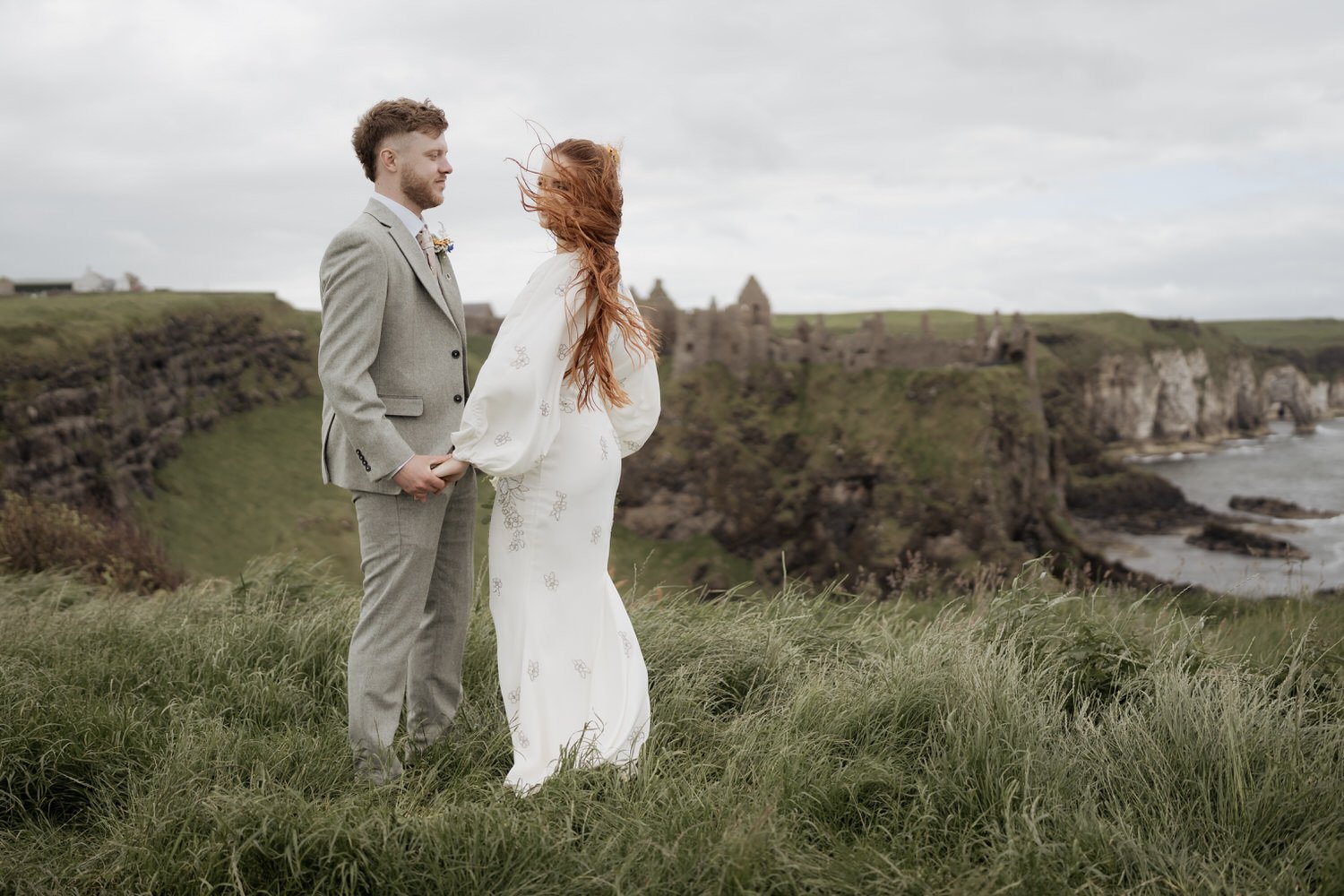 Bride and groom standing together on a coastal clifftop with wind-blown hair and overcast skies, embracing the real moment of their wedding day
