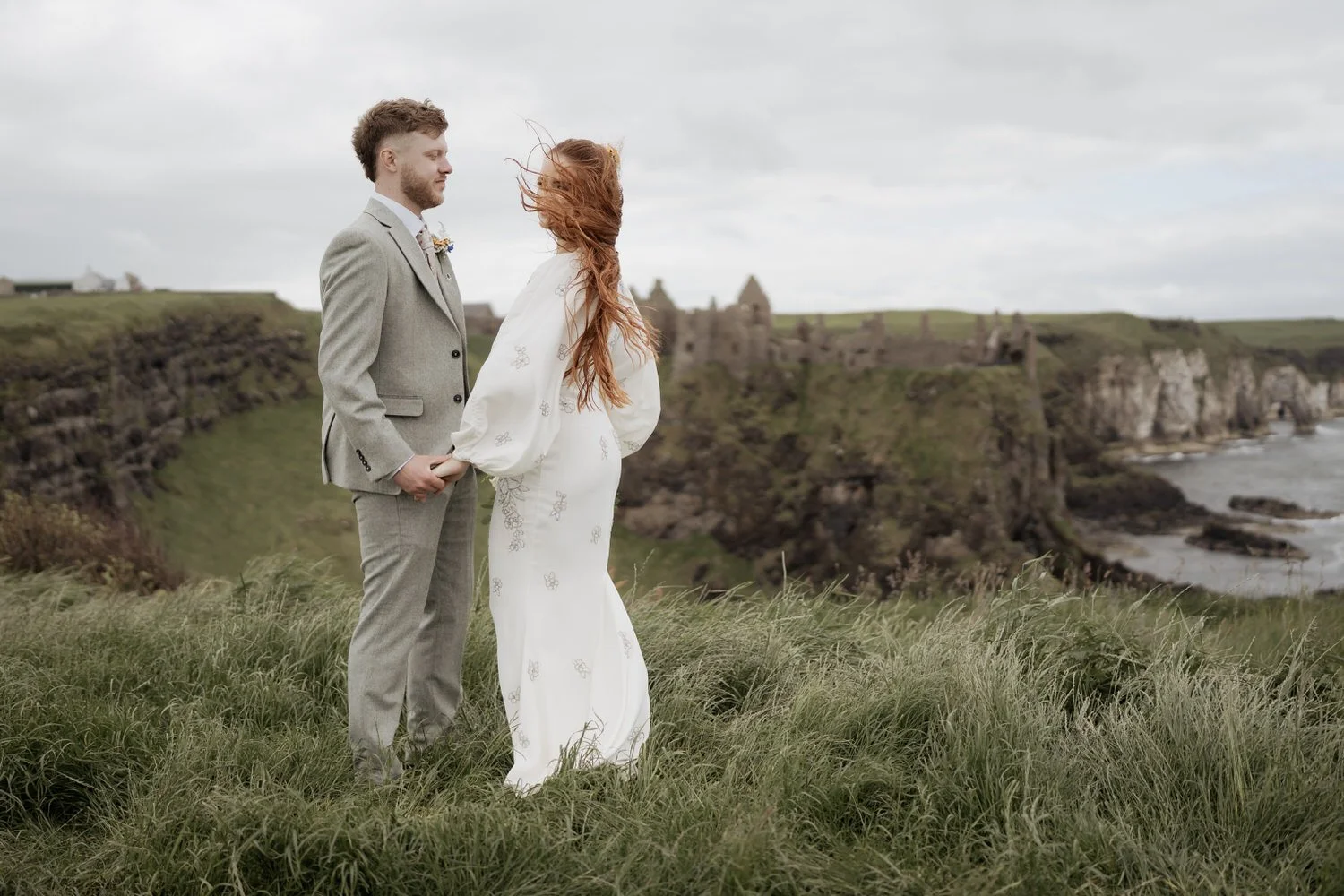 Bride and groom standing together on a coastal clifftop with wind-blown hair and overcast skies, embracing the real moment of their wedding day