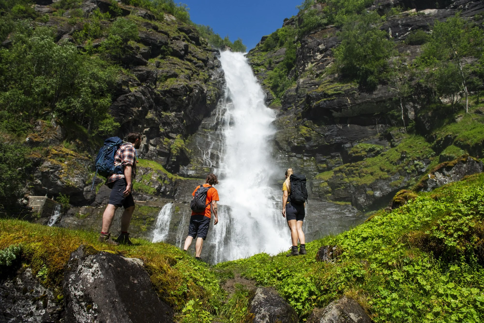 Three hikers with backpacks standing near a waterfall in a lush green forest, with a clear blue sky above.