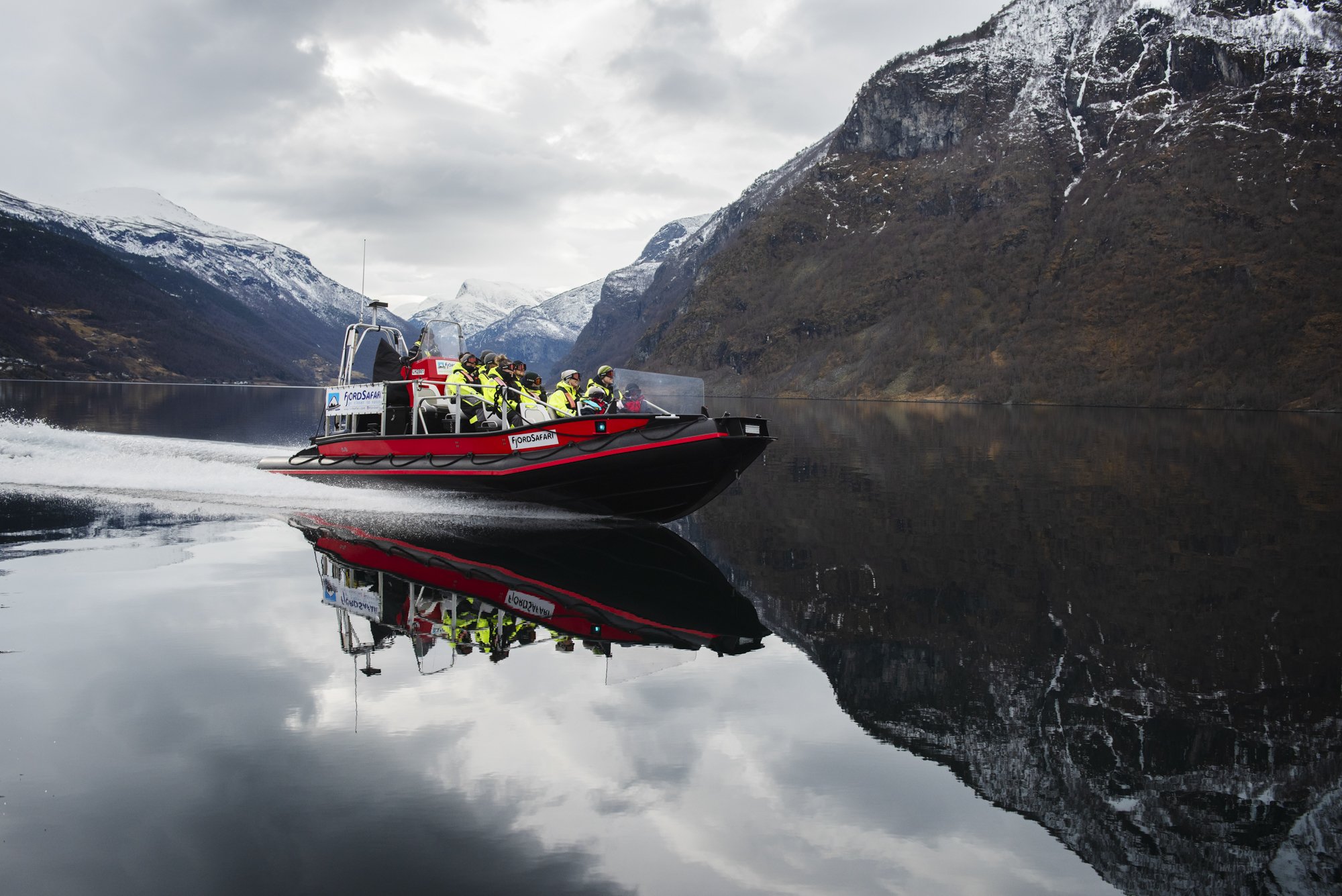 Rød og svart åpen RIB-båt med folk iført flytedrakter og googles. Vannet er veldig stille og ser ut som et speil. Store snødekte fjell i bakgrunnen.