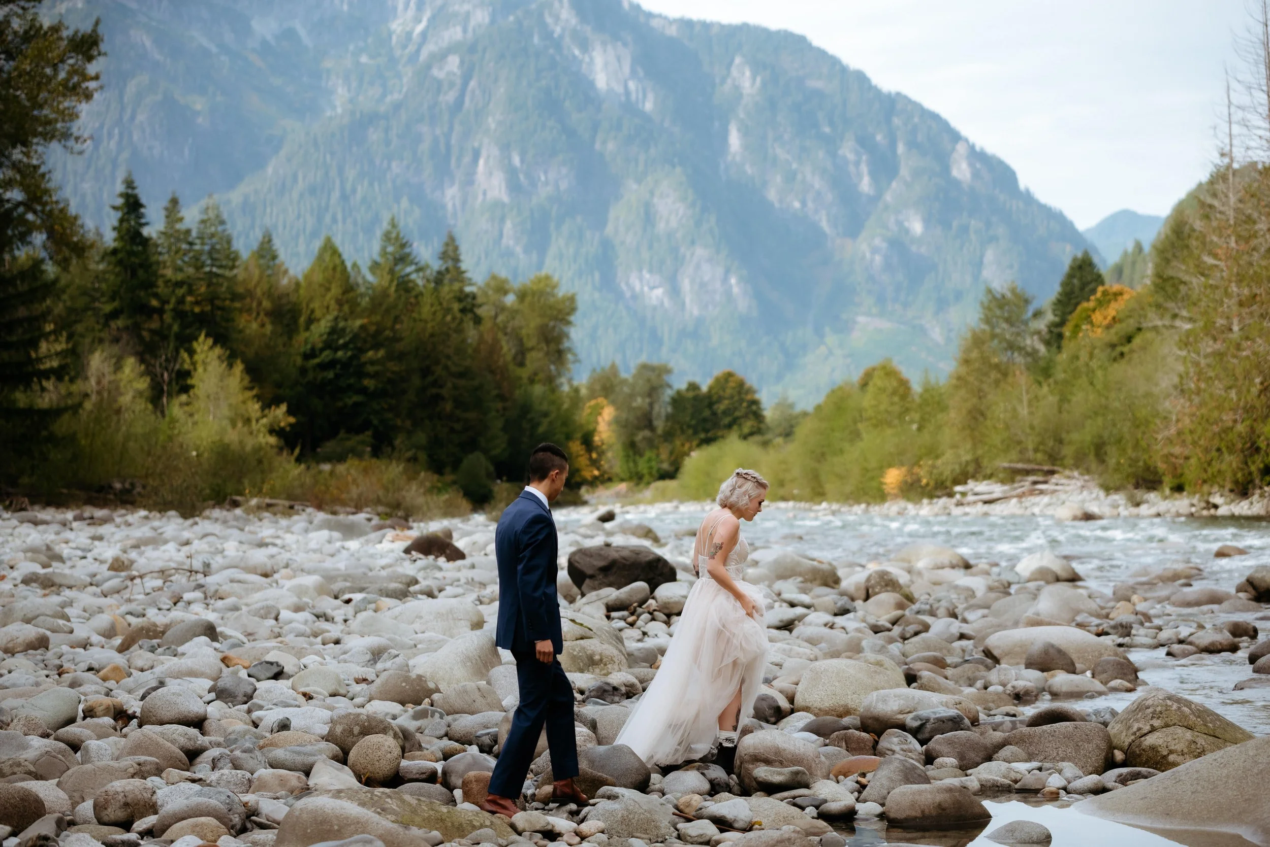 A bride and groom hike over river rocks in Index, Washington during their elopement