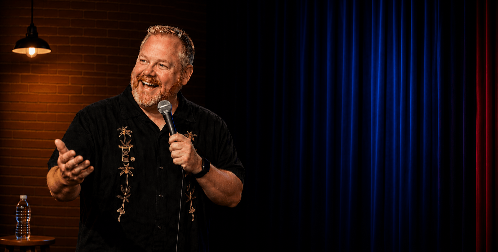 A man with gray hair and a beard, wearing a black shirt with embroidered designs, holding a microphone and smiling while performing on stage with a dark blue curtain background and a brick wall with a hanging lamp behind him.