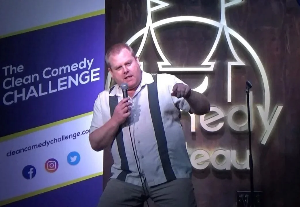 SK Morton performing stand-up comedy at 'The Comedy Challenge.' He is holding a microphone and gesturing with his other hand. There are neon signs and a banner with social media icons in the background.