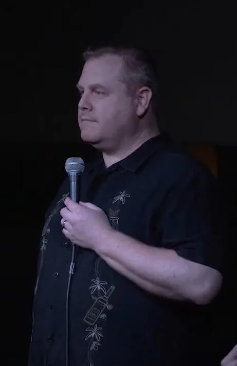 SK Morton holding a microphone during a stand-up comedy performance at Sally's Tomatoes, wearing a black shirt with palm tree embroidery.