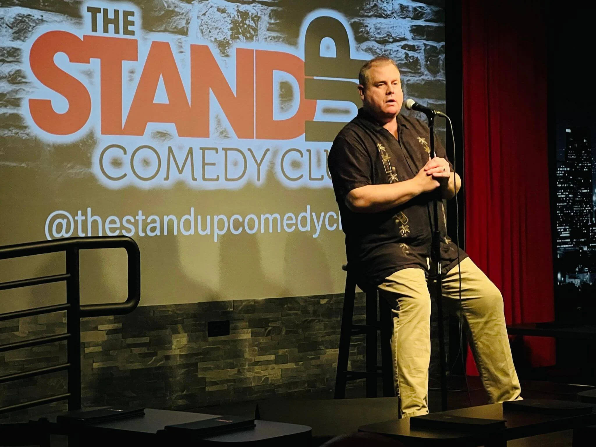 SK Morton sitting on a stool speaking into a microphone on a stage, with a backdrop displaying a logo for 'The Stand-Up Comedy' and a cityscape at night.
