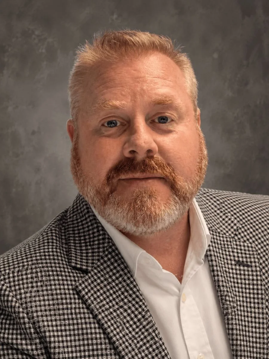 Professional headshot of SK Morton with a full beard wearing a black-and-white checkered blazer and white shirt against a gray background.