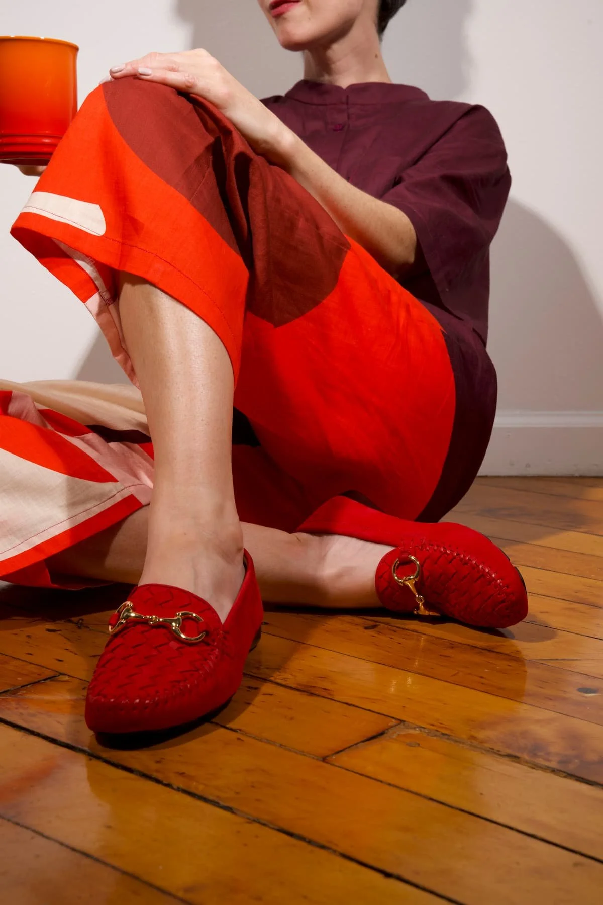 A woman in a maroon dress and red shoes with gold chain details is seated on a wooden floor, resting her right hand on her left knee, with a red and orange fabric in her lap, and a red vase nearby.