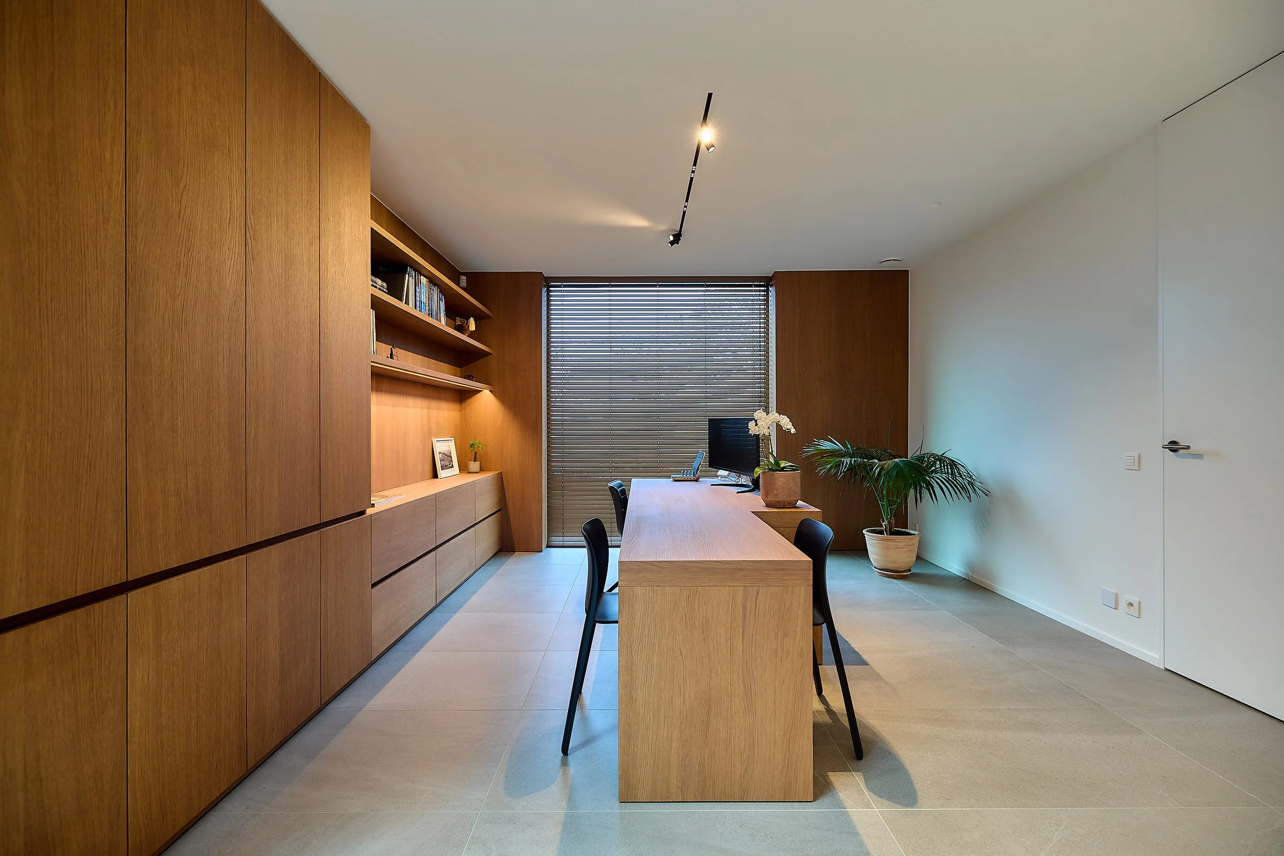 Modern kantoorruimte met houten meubels, planten, en grote ramen met jaloezieën.