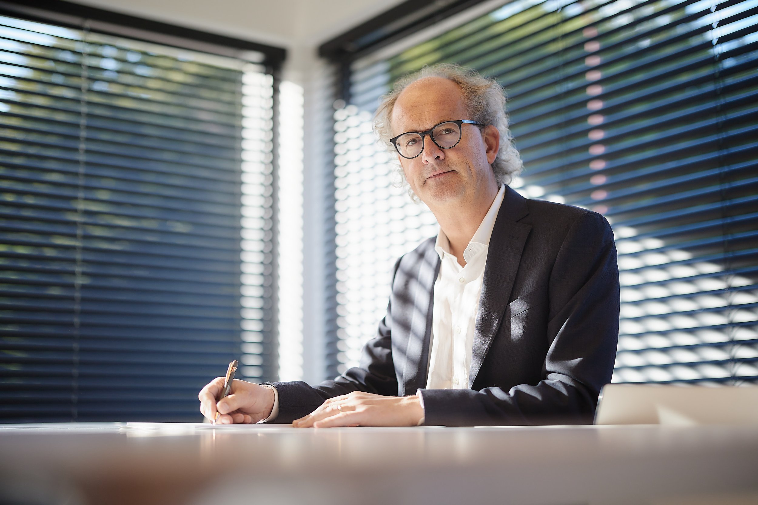 Een oudere man met grijs, golvend haar en brillen, gekleed in een zwart pak en witte overhemd, zit aan een bureau met donkere jaloezieën op de achtergrond.