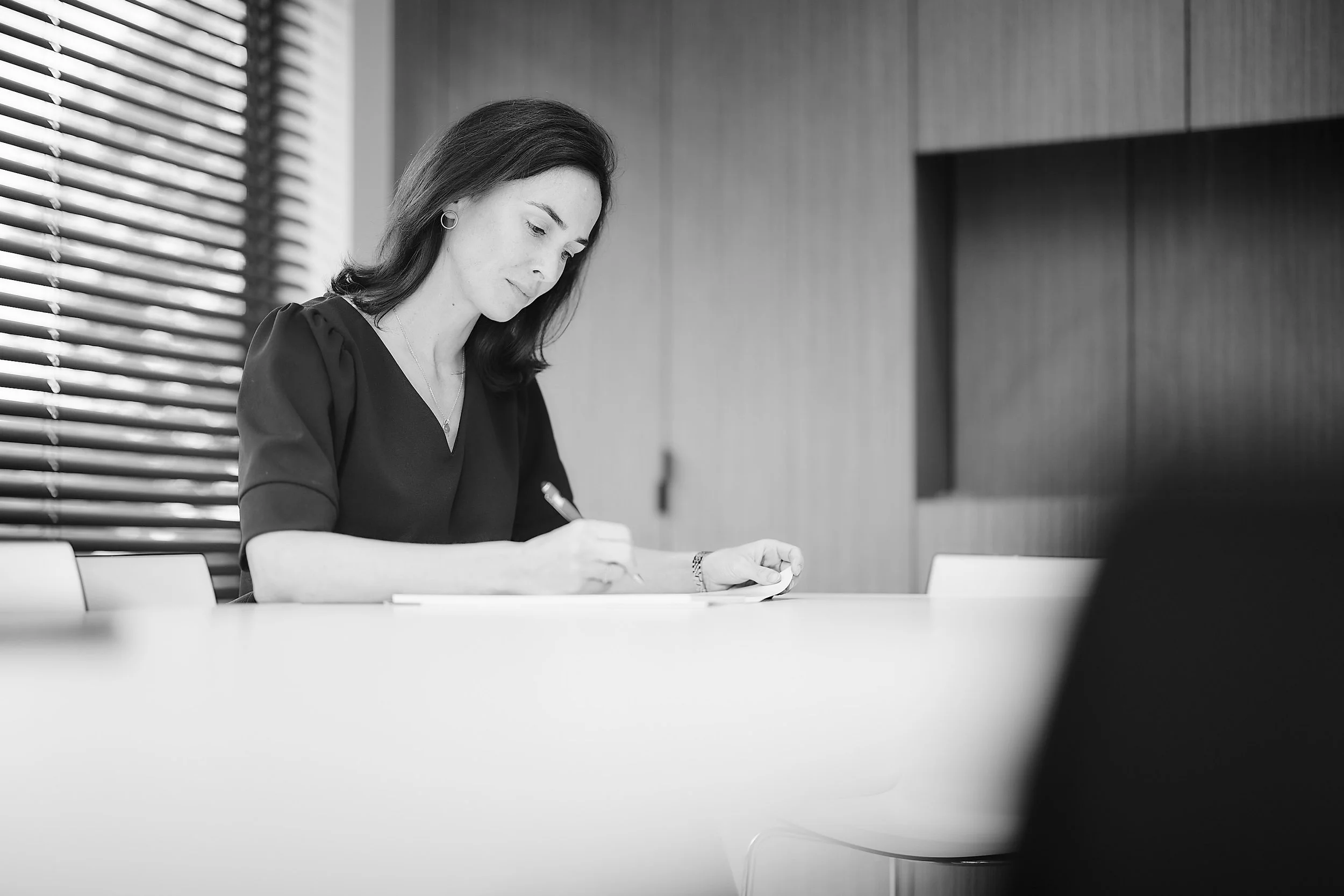 Een vrouw schrijft notities tijdens een vergadering in een kantoor met houten wanden en een raam met jaloezieën.