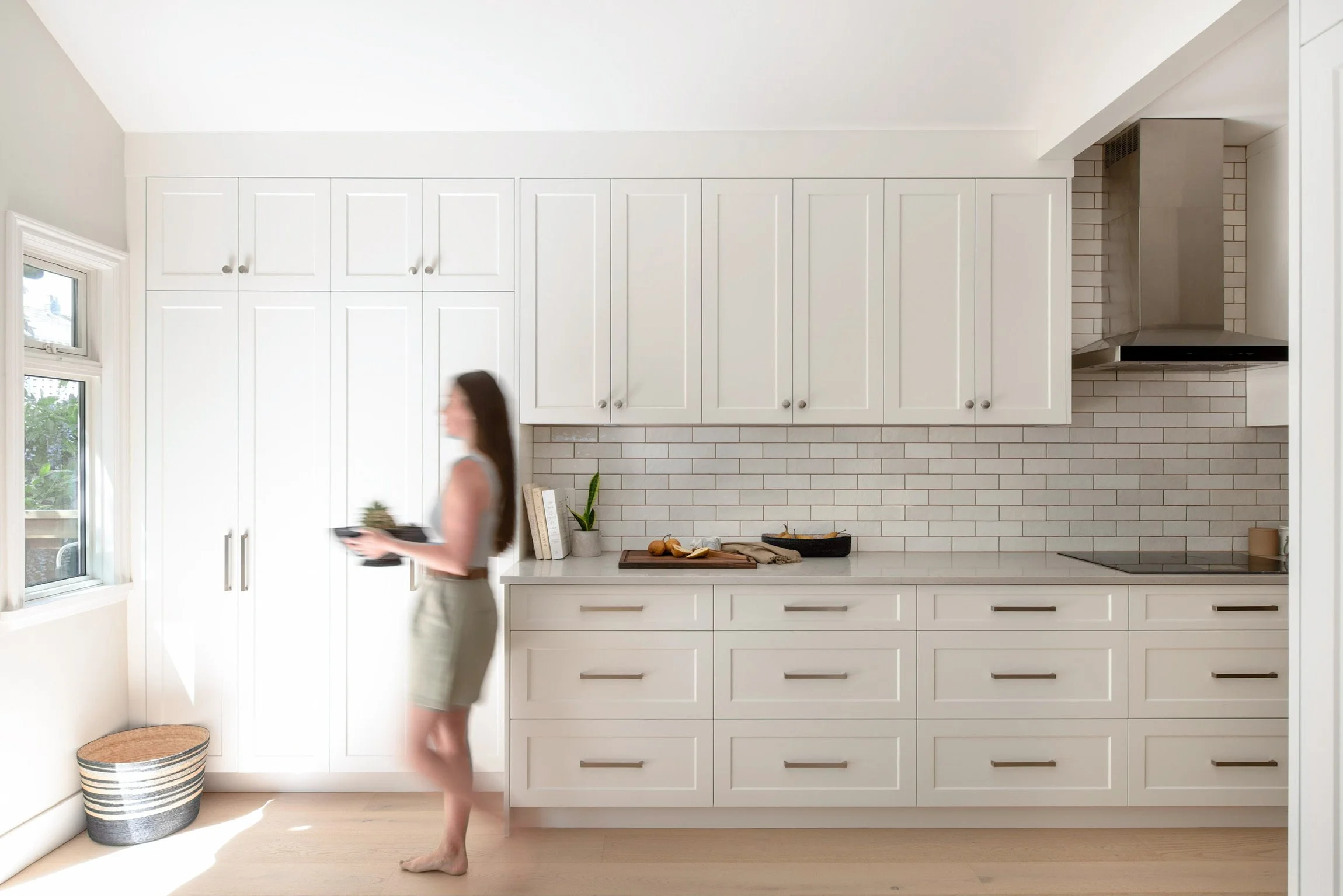 Bright kitchen with white cabinets, a woman walking with a tray, a window, and a basket on the floor. Architectural interior photographer, Vancouver bc