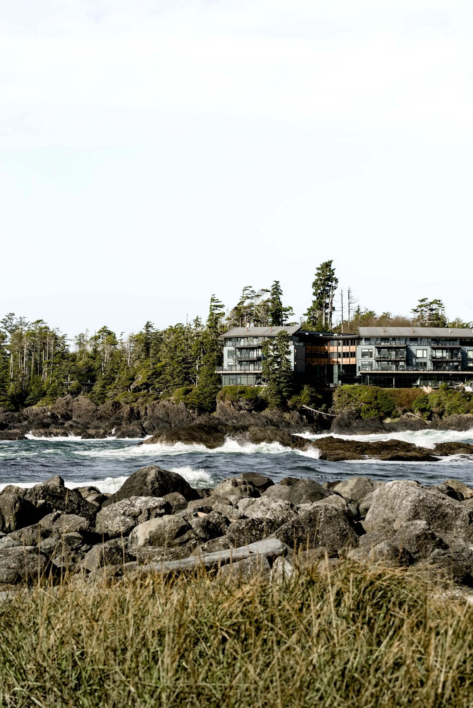 Coastal scene featuring rocks and waves in the foreground, with modern multi-story apartment buildings surrounded by trees on a hillside in the background.