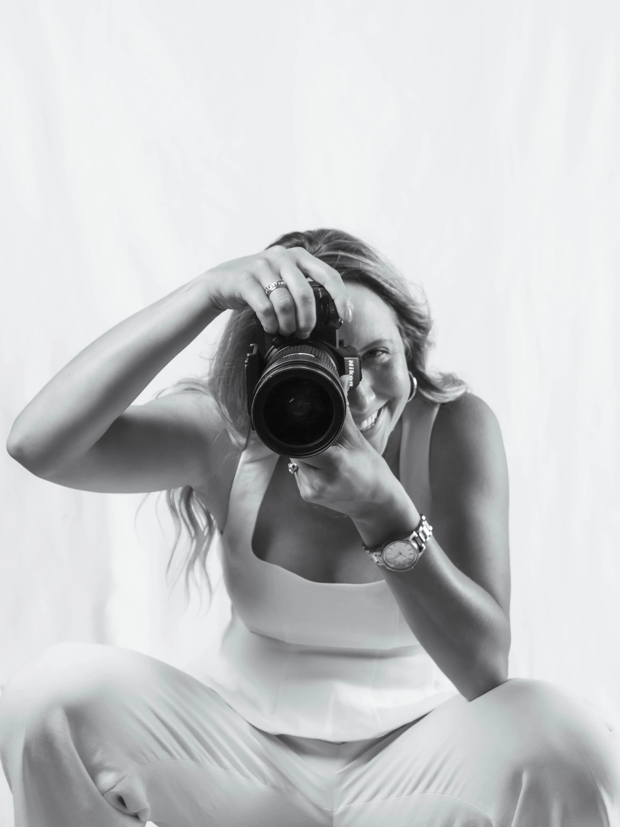 Photographer with long hair, wearing a sleeveless top and pants, taking a  photograph with a Nikon mirrorless camera in front of a plain background.