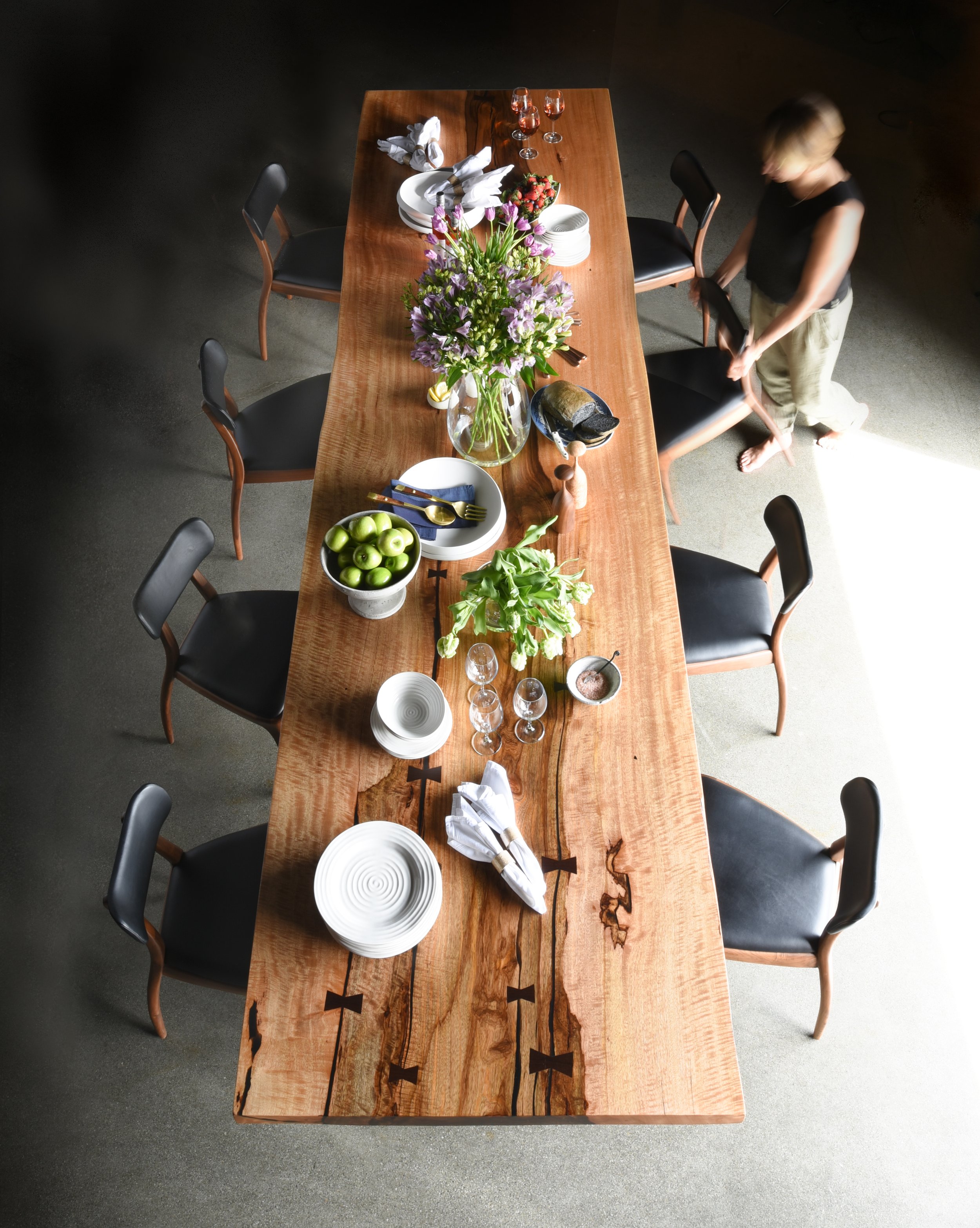 product photography of a styled luxury wooden dining table Birds Eye view and a woman walking into the scene with the table is set and there are chairs set up around it. shot for Reduxwood West