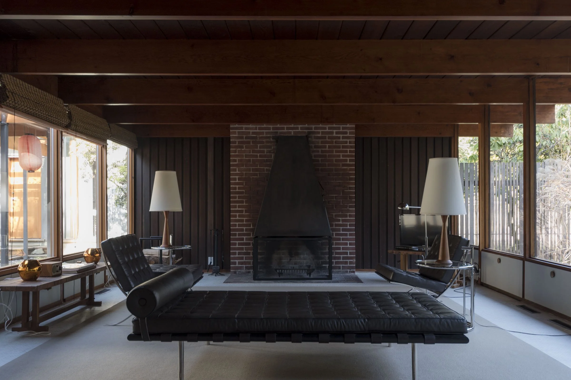 Living room with a brick fireplace, black leather chaise lounge, two table lamps, a small TV on a stand, and large windows with blinds.