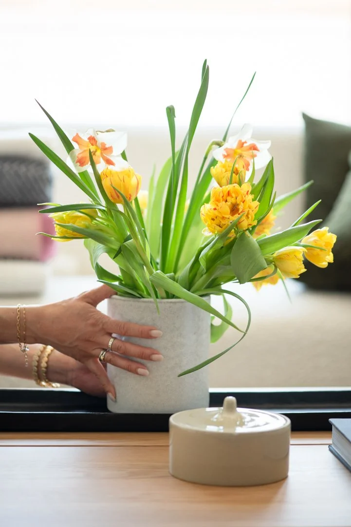 product photography of a woman's hands putting a vase of yellow tulips on a white oak table