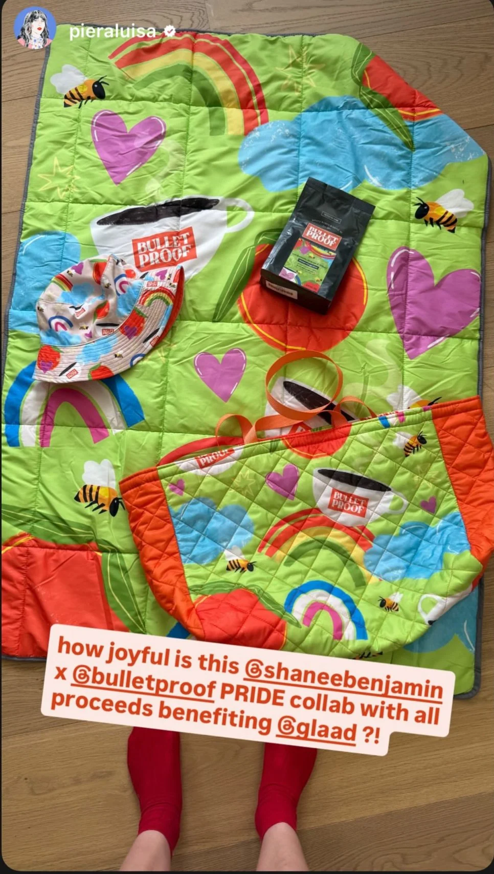 Colorful quilt with rainbow, blue clouds, pink hearts, and bees, spread out on a wooden floor. Items on the quilt include a patterned hat, a black Bulletproof coffee packet, a pair of glasses, and a tote bag with matching rainbow and bee design. A person wearing red socks stands at the bottom of the quilt. Text overlay reads: 'how joyful is this @shanebenjamin x @bulletproof PRIDE collab with all proceeds benefiting @glaad?!'