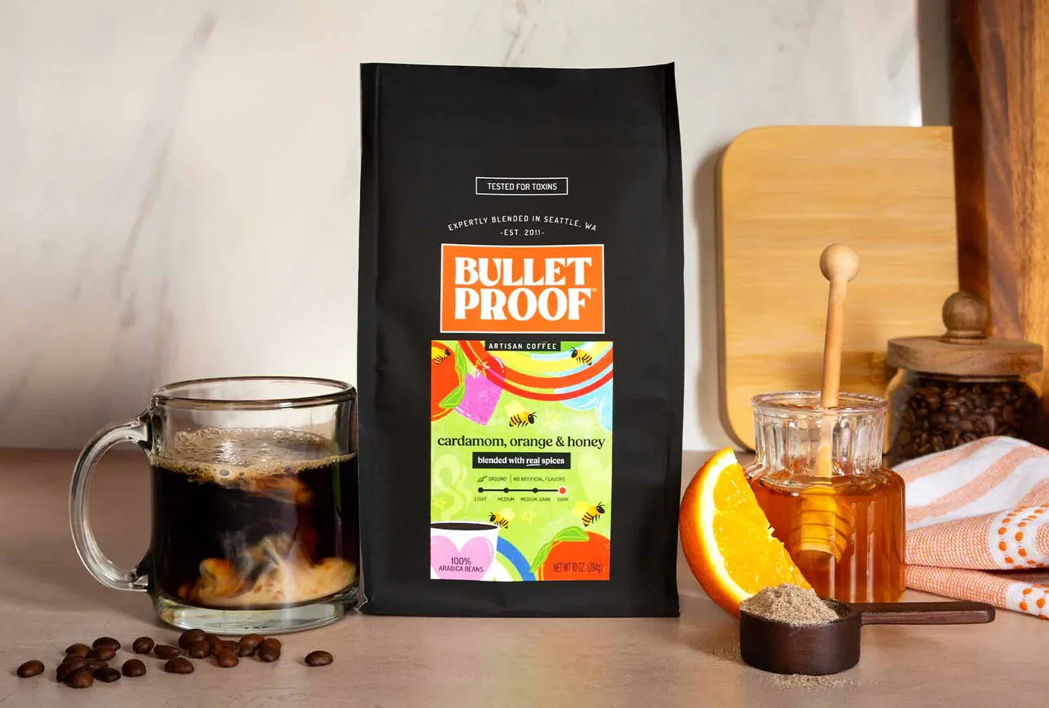 A package of Bulletproof brand coffee labeled "cardamom, orange & honey" standing on a light-colored surface. To the left, a glass mug filled with black coffee, and below it, scattered coffee beans. To the right, a small jar of honey with a honey dipper inside, a wedge of orange, and a small dish of ground spices. In the background, wooden cutting boards and a glass jar with coffee beans. There is a striped cloth on the right side of the image.