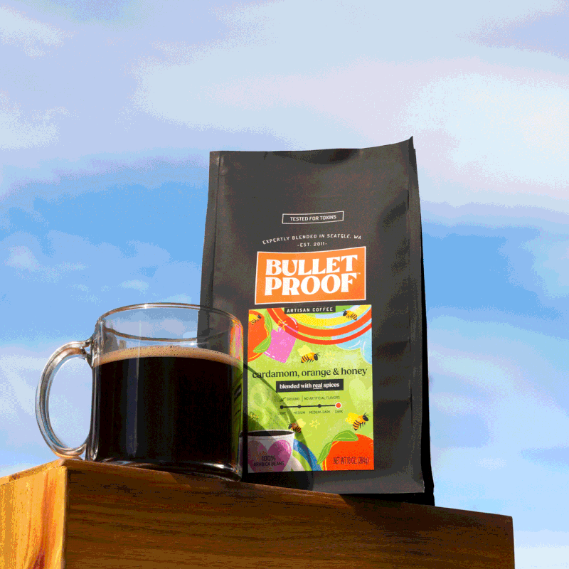 A black bag of Bulletproof artisan coffee with cardamom, orange, and honey flavor, placed on a wooden surface next to a clear glass mug filled with dark coffee, against a background of blue sky with some clouds.