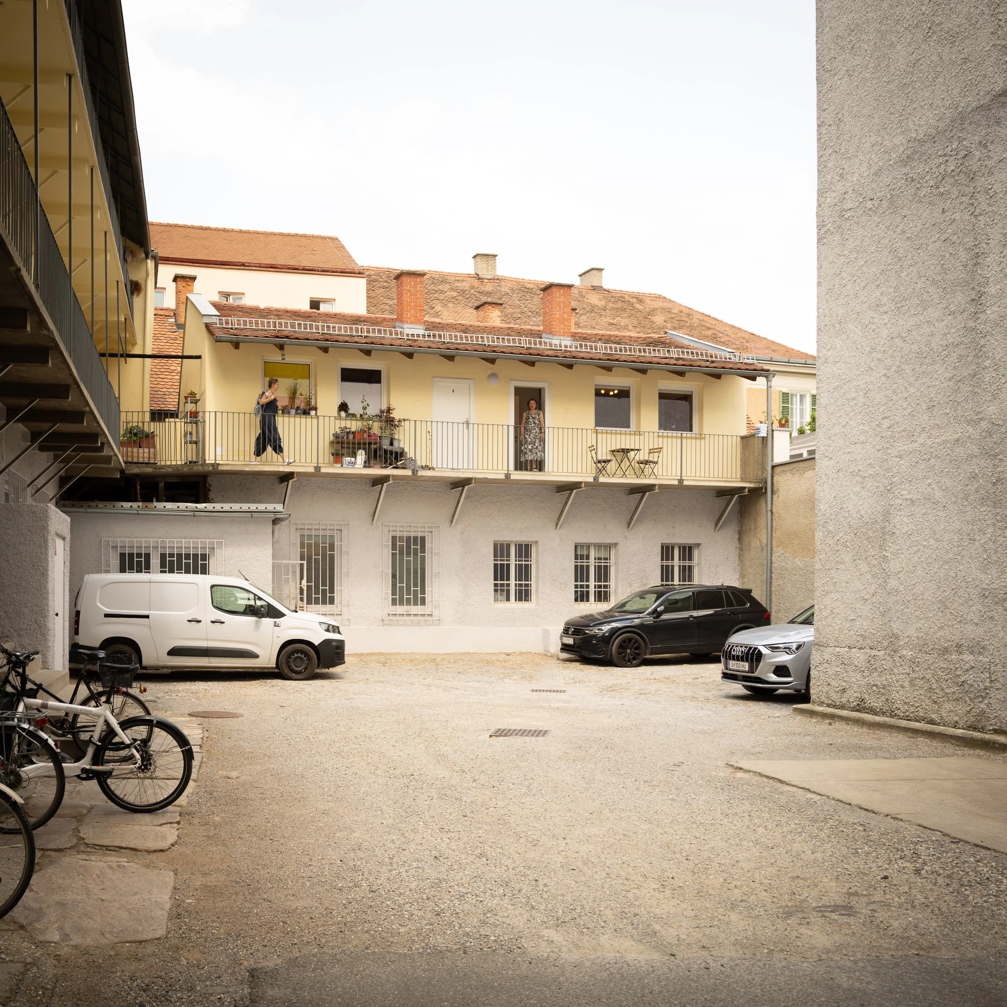 Innenhof mit Parkplätzen vor Wohnhaus mit Balkon und Dachfenster, zwei Frauen sprechen miteinander.