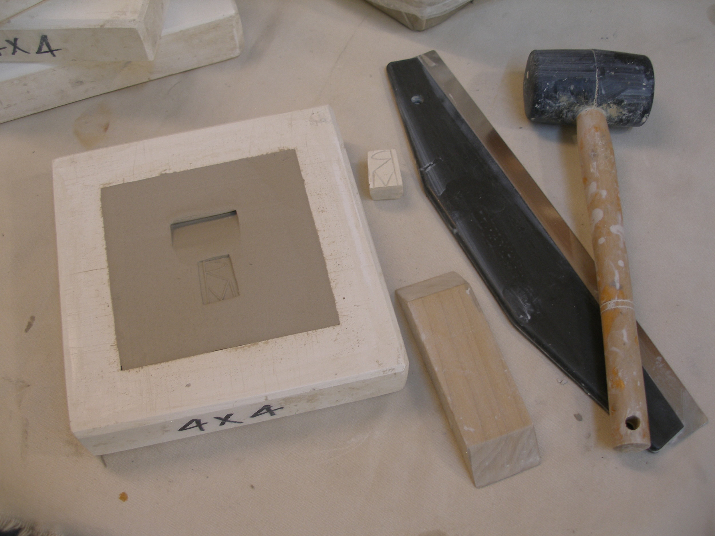 The excess clay is sliced off with a wire tool, and the back of the tile is scraped and smoothed flat. A beveled piece of wood is pressed in to create an indentation for hanging. The tile is also stamped with the maker's mark. The tile will dry for several hours. As the plaster draws moisture from the clay, the tile begins to shrink away from the mold. The mold can then be inverted onto a board, the tile will drop out, and the mold removed.